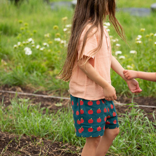 Child wearing a peach shirt and blue shorts with red apples in a garden setting
