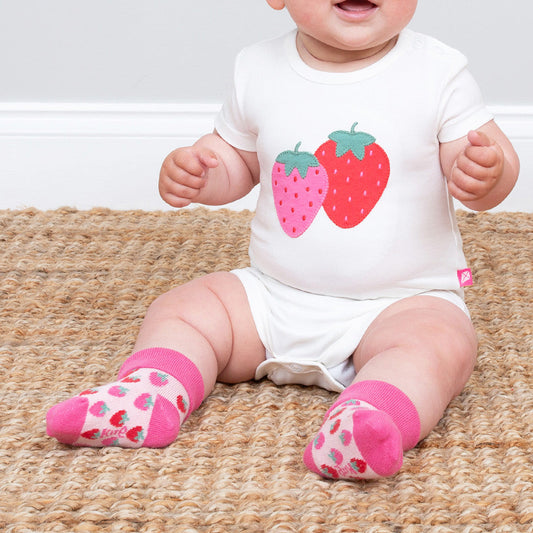 Baby wearing a onesie with strawberry design and pink socks on a beige rug.