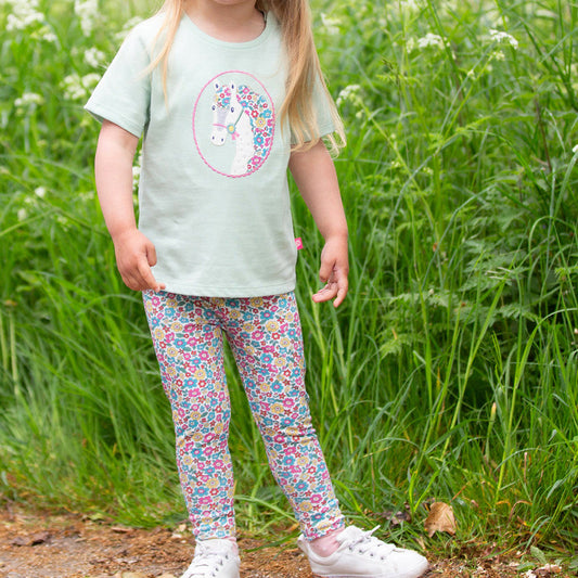 Child wearing a light green t-shirt with a floral unicorn design and colorful leggings standing in a field of tall grass.