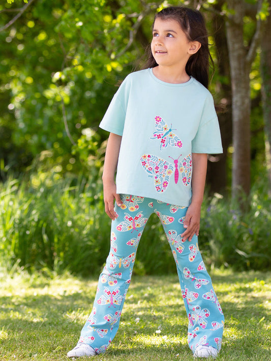 Young girl wearing a light blue t-shirt and butterfly leggings standing outdoors.