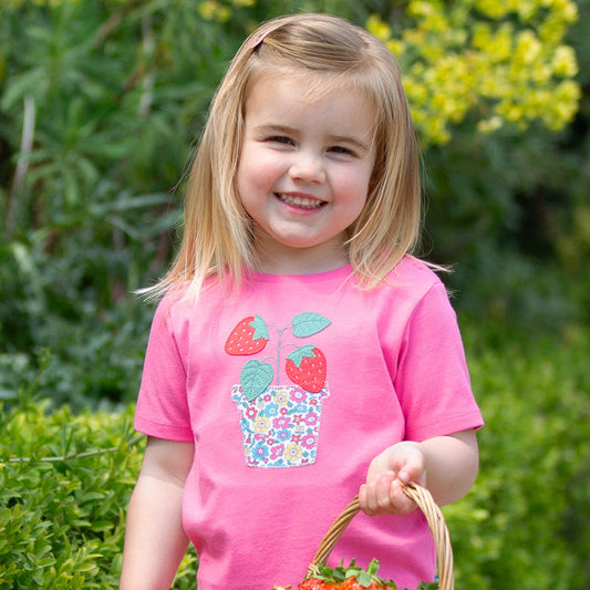 Young girl in a pink shirt with strawberry design holding a basket of strawberries outdoors.