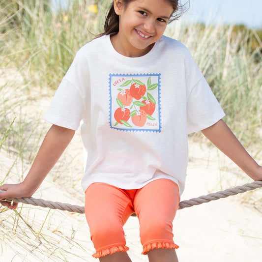 Young girl sitting on a rope swing at the beach wearing a white t-shirt with a colorful design and orange shorts.