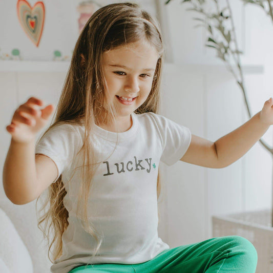 Young girl wearing a white t-shirt with 'lucky' printed on it, sitting indoors.