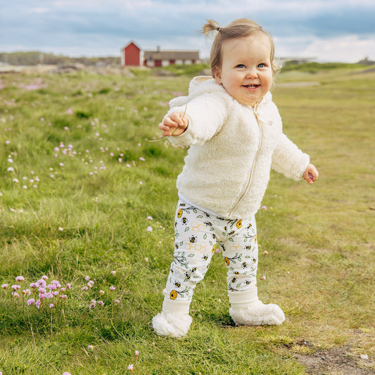 Child in a white fluffy coat and bumblebee patterned pants standing in a grassy field with a red building in the background.