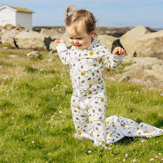 Child in a bumblebee onesie standing in a grassy field with flowers and rocks in the background