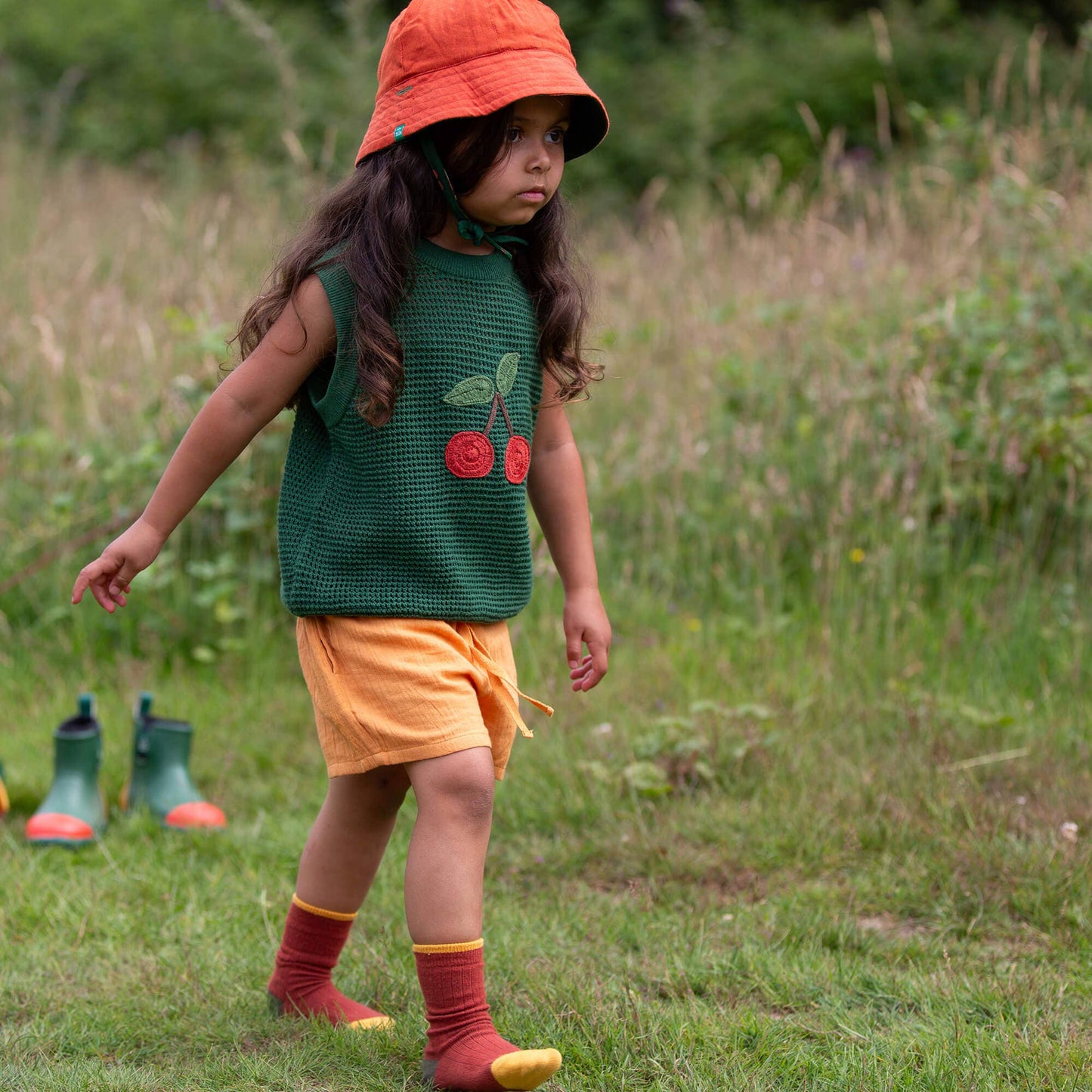 Child wearing a green sleeveless vest with cherry design, orange shorts, and red socks standing in a grassy field.