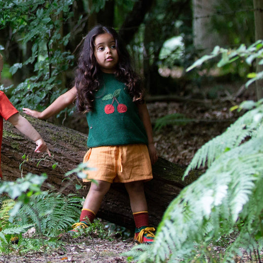 Child in a forest wearing a green shirt with red cherries and orange shorts.
