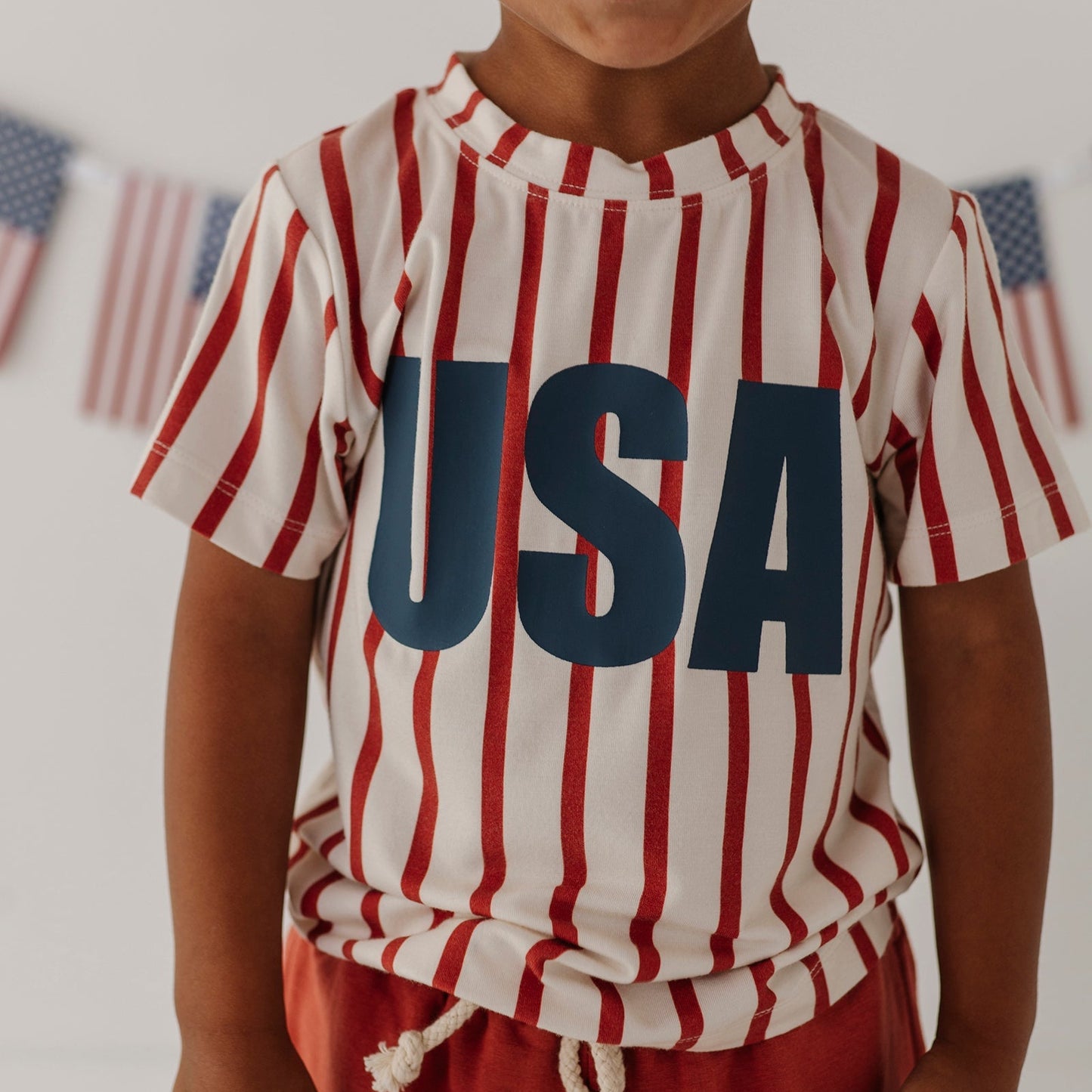 Child wearing a red, white, and blue striped shirt with 'USA' printed on it, standing in front of American flags.