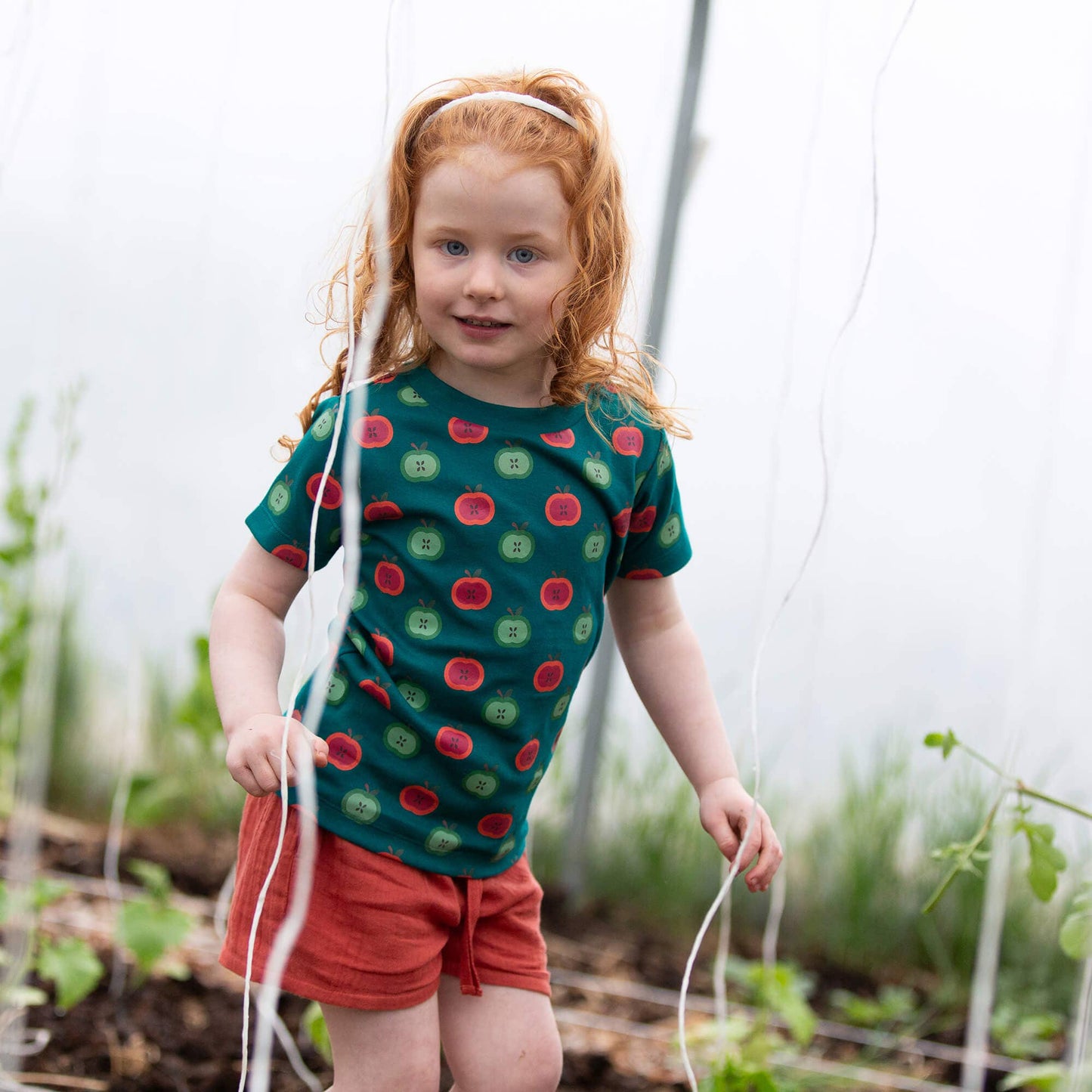 Young girl in a garden wearing a green shirt with red and green pattern and red shorts.