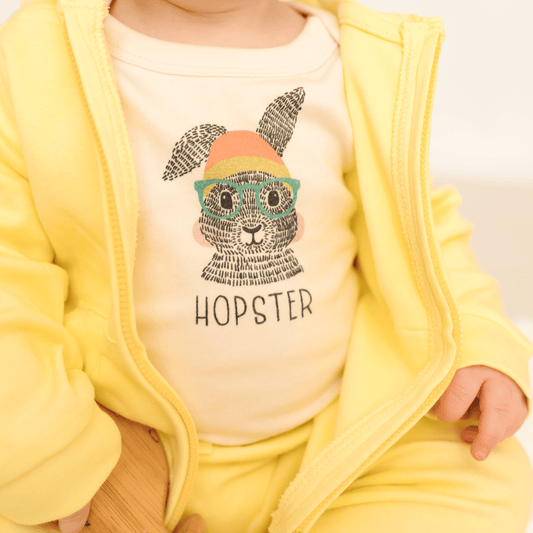 Baby wearing a yellow onesie with a graphic of a rabbit wearing a hat and sunglasses, and the word 'Hopster'.