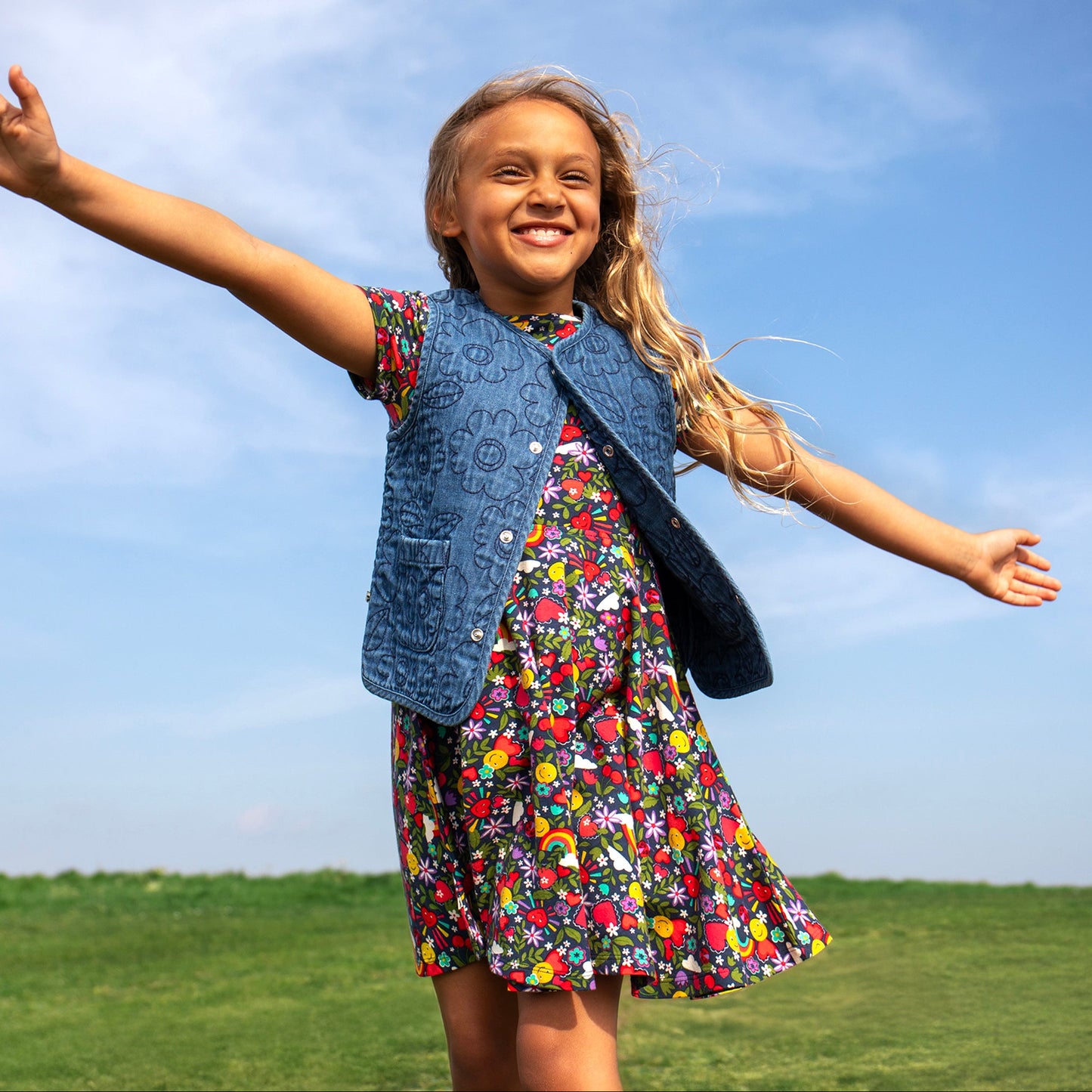 Young girl in a colorful dress and denim vest standing on a grassy field with a blue sky.