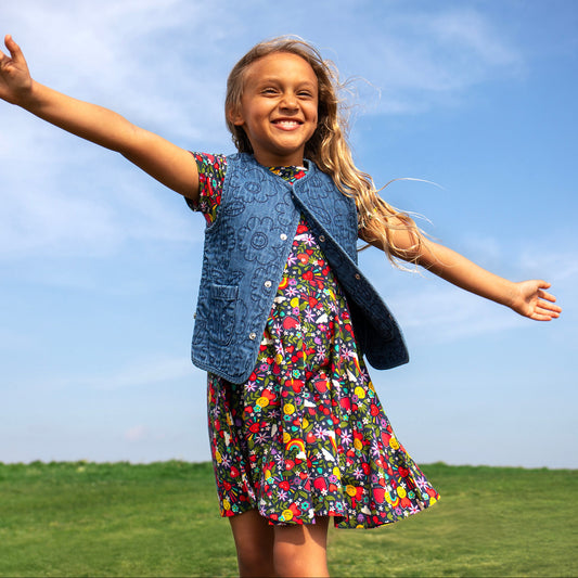 Young girl in a colorful dress and denim vest standing on a grassy field with a blue sky.