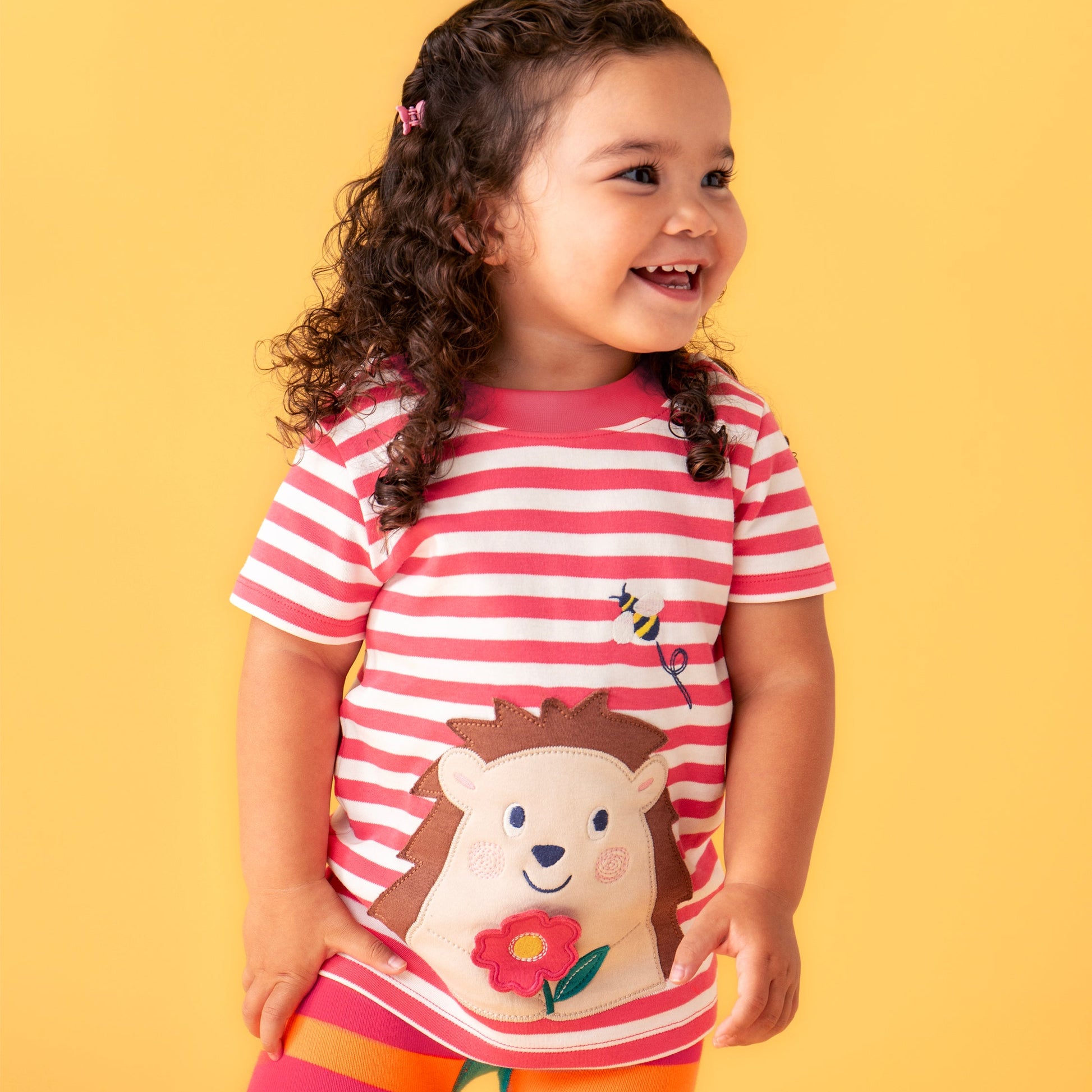 Child wearing a pink and white striped shirt with a hedgehog design and orange and pink striped pants on a yellow background