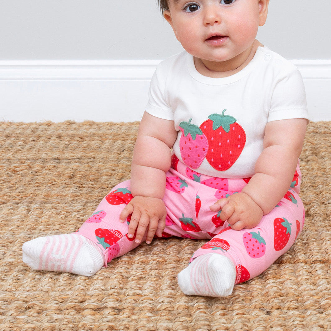 Baby wearing a white shirt with strawberry design and pink strawberry print pants sitting on a woven mat.