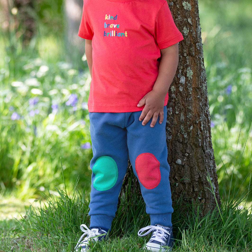 Child wearing a red shirt and blue pants with colorful patches standing next to a tree in a grassy area.