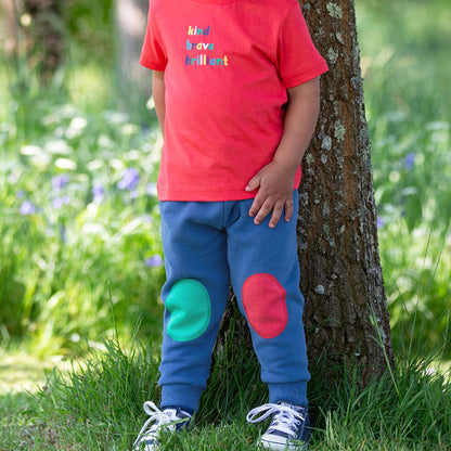 Child wearing a red shirt and blue pants with colorful patches standing next to a tree in a grassy area.