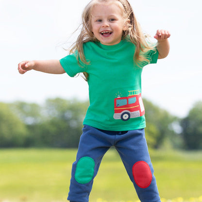 Child wearing a green shirt with a fire truck design and blue pants with knee pads, jumping outdoors.