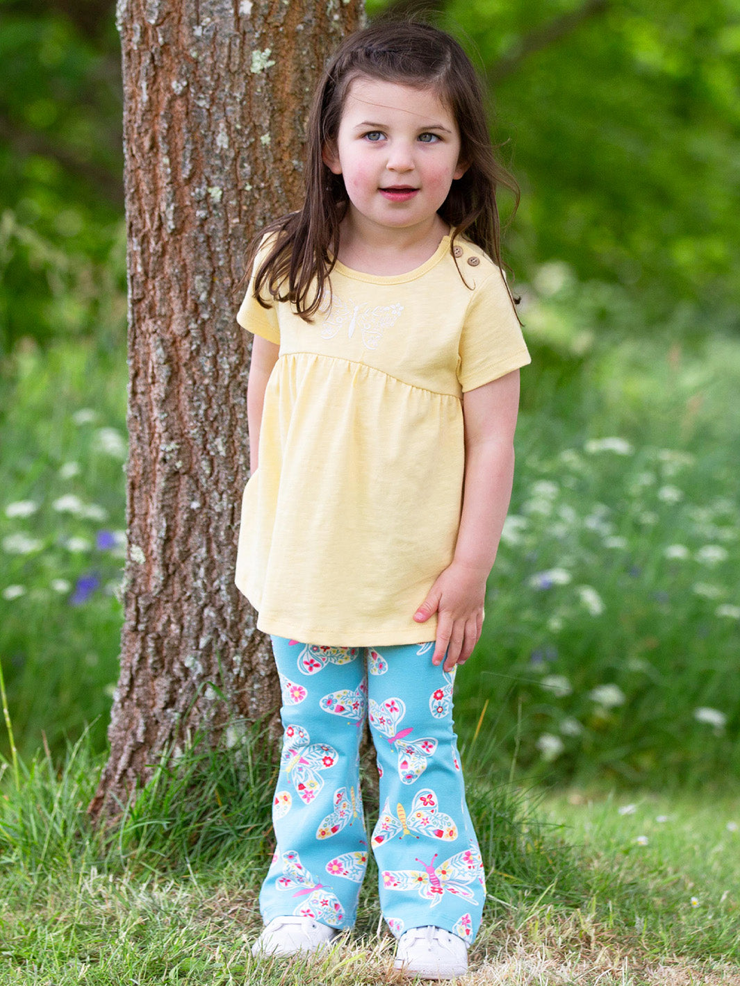 Young girl in a yellow top and blue butterfly pants standing next to a tree in a grassy area with flowers.