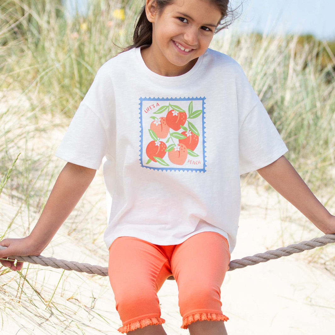 Young girl sitting on a rope swing at the beach wearing a white t-shirt with a colorful design and orange shorts.