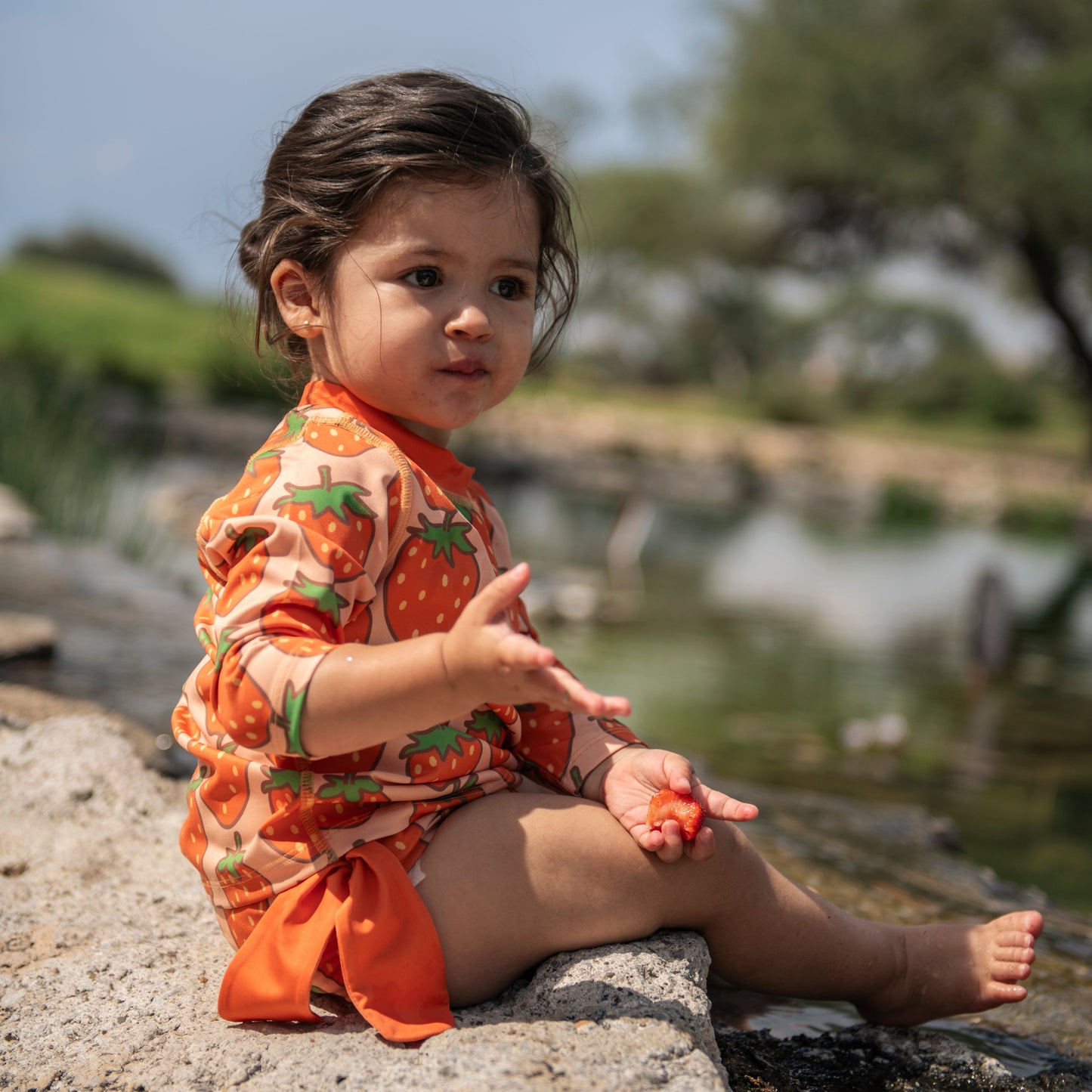 Child in an orange outfit sitting on rocks by a body of water with trees in the background