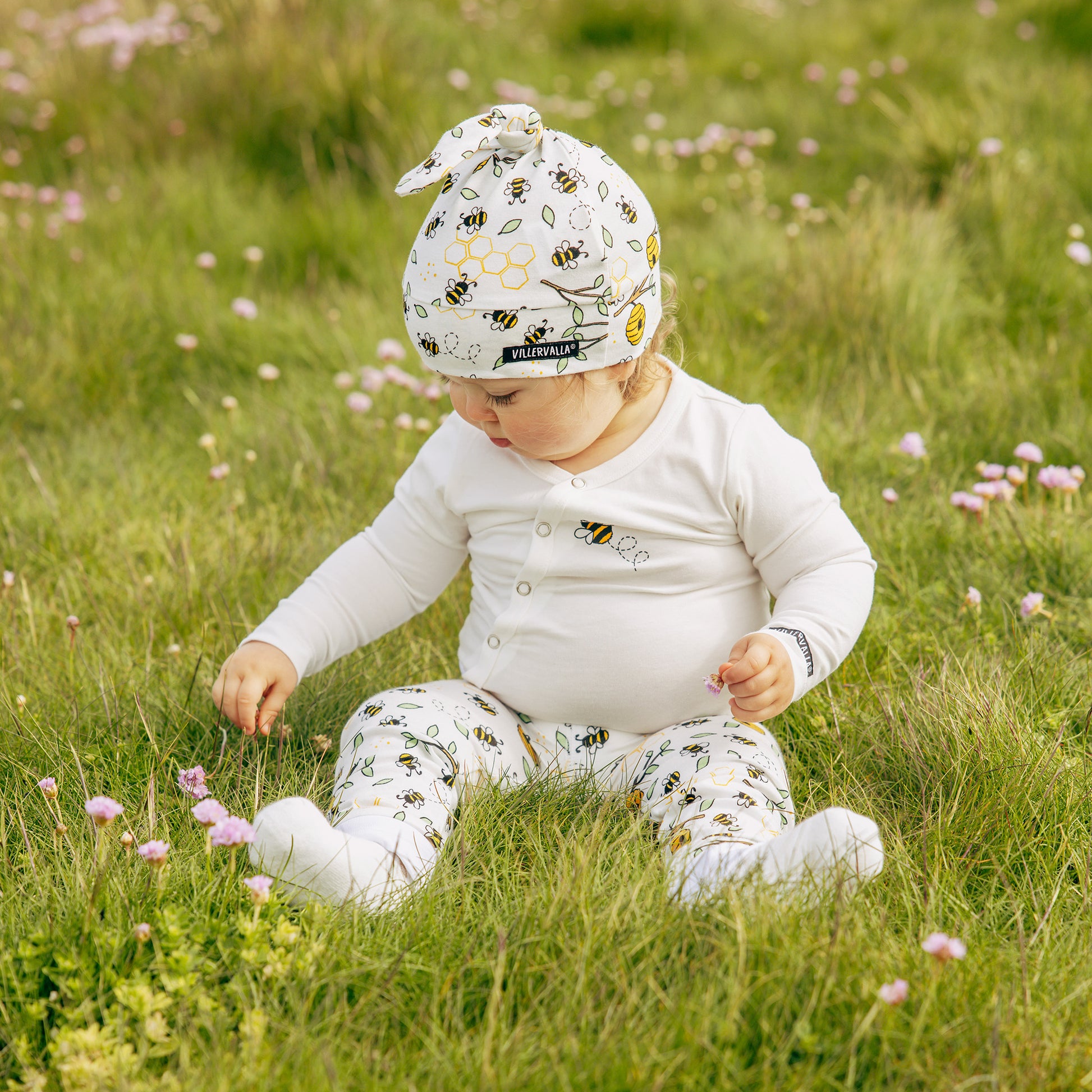Child sitting in a grassy field wearing a white outfit with bee patterns and a matching hat.
