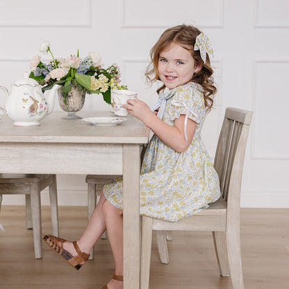 Young girl in a floral dress sitting at a table with a teacup, surrounded by flowers and a teapot.