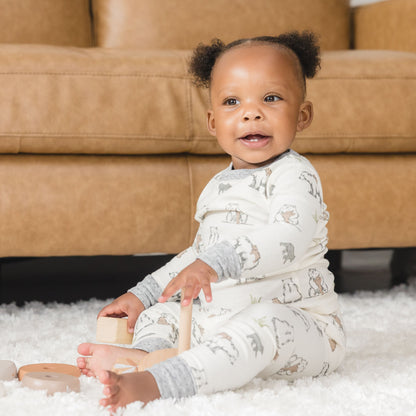 Baby sitting on a carpeted floor holding wooden toys, wearing a patterned onesie in front of a brown couch.