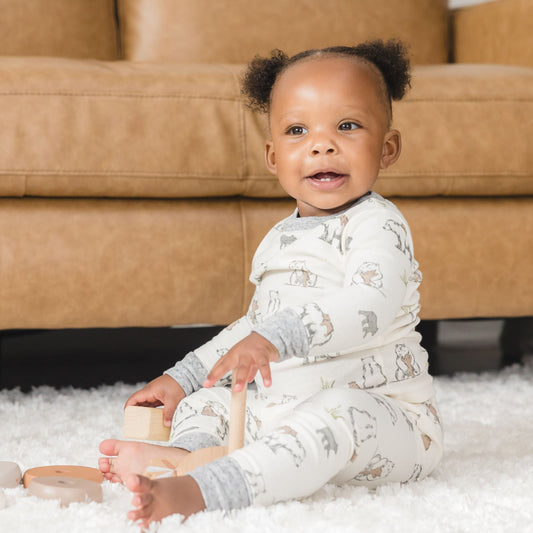 Baby sitting on a carpeted floor holding wooden toys, wearing a patterned onesie in front of a brown couch.