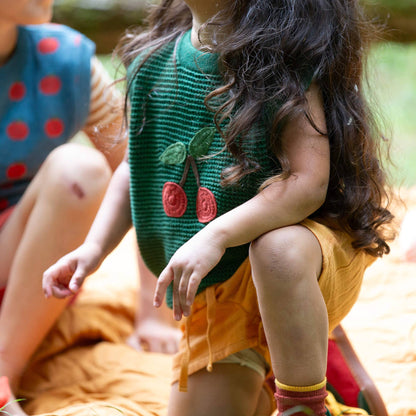 Child wearing a green knitted sweater with cherry design sitting outdoors.